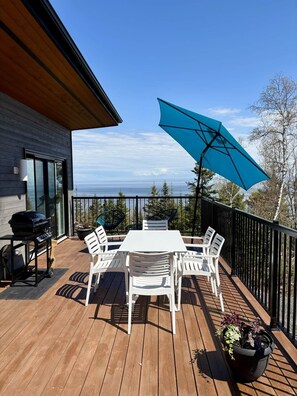 Une terrasse en bois avec une table et des chaises blanches, un barbecue et un parasol bleu.