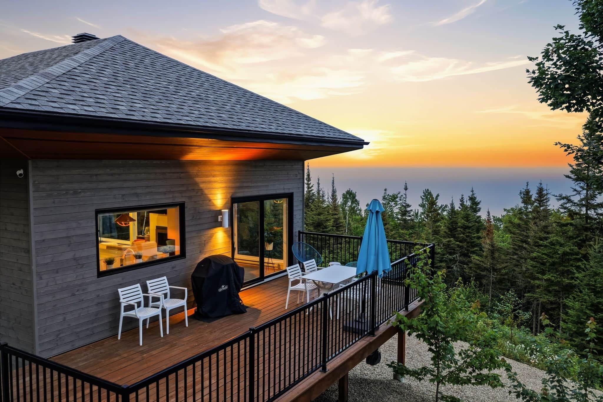 A modern house with a deck, large windows, and a view of the ocean at sunset.