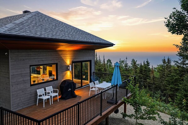 Une maison moderne dotée d’une terrasse, de grandes fenêtres et offrant une vue sur l’océan au coucher du soleil.