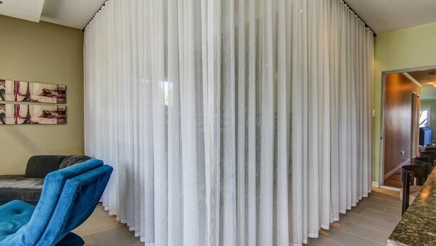 A room with a large white curtain partition, a blue armchair, a dining area with a marble table, and a painting on the wall.