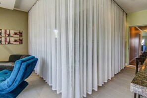 Una habitación con un amplio separador de tela blanca, un sillón azul, un comedor con mesa de mármol y un cuadro en la pared.