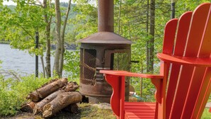 Une chaise Adirondack rouge est placée à côté d’un foyer en métal contenant des bûches, le tout se dressant sur fond de lac et d’arbres.