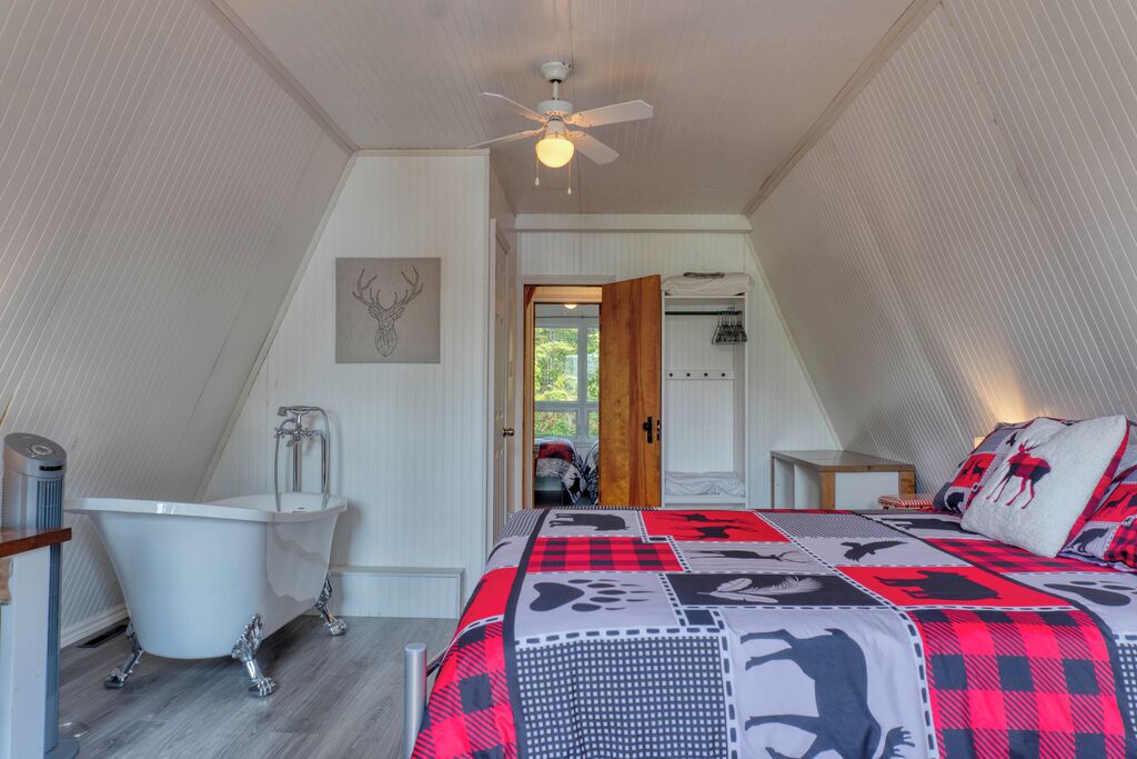 Ein Schlafzimmer mit schrägem Dach, einer weißen Badewanne, einem Bett mit rot-schwarz gemustertem Überwurf und einem Deckenventilator.