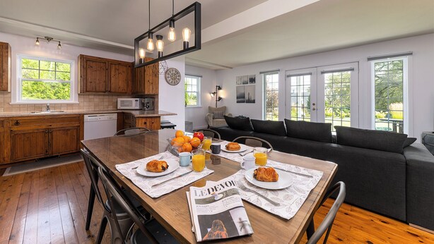 Una cocina moderna con armarios de madera, una mesa de comedor lista para el desayuno y un área de estar con sofá y ventanas amplias.