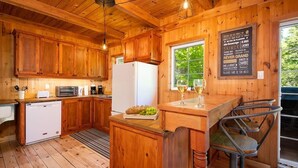 A wooden cabin-style kitchen with wooden cabinets, a white refrigerator, a microwave, and a sink. There is a window with a view of greenery outside, and a chalkboard menu on the wall.