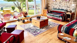 Ein gemütliches Wohnzimmer mit einer Steinwand, einem farbenfrohen Sofa, einem Holztisch und Blick auf das Meer durch große Fenster.