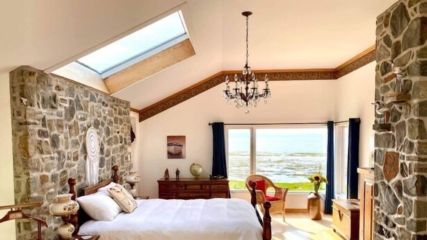 Ein Schlafzimmer mit Steinwand, einem hölzernen Bett, einer Chandelüster, einem Schminktisch und Meerblick durch große Fenster.