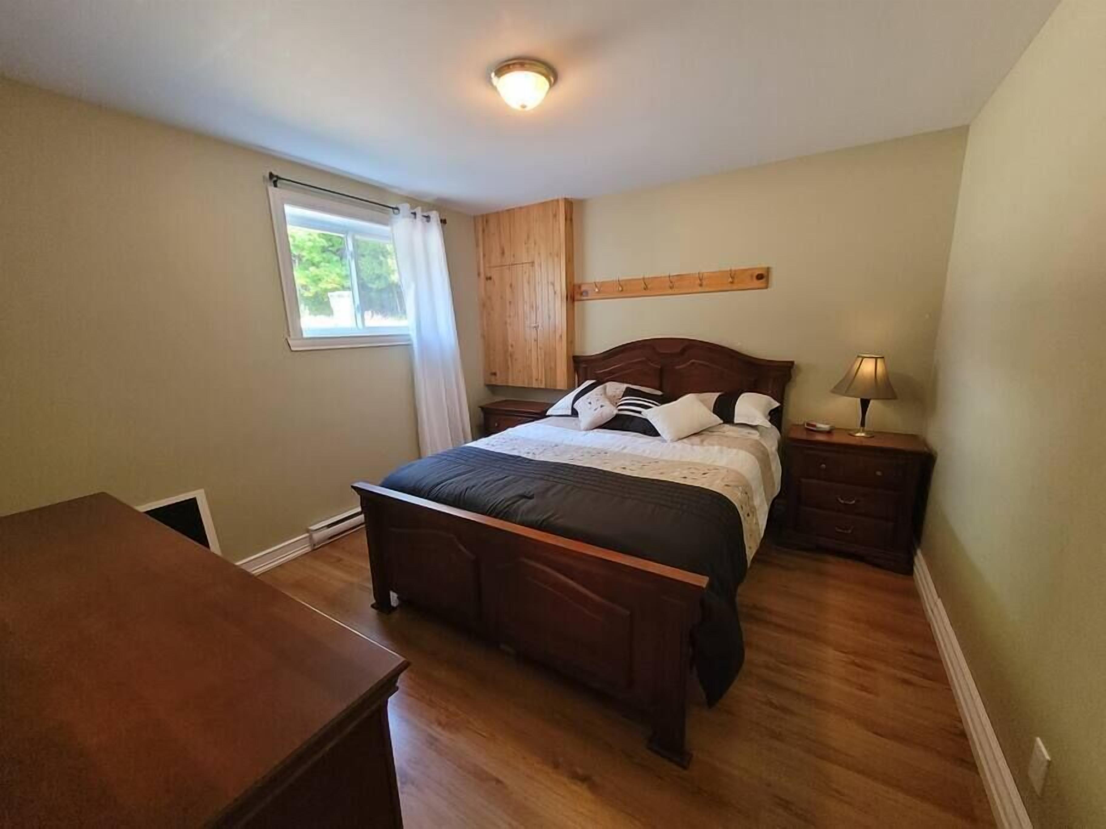 A bedroom with a wooden bed, a nightstand with a lamp, a window with curtains, and a wooden dresser.