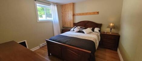 A bedroom with a wooden bed, a nightstand with a lamp, a window with curtains, and a wooden dresser.