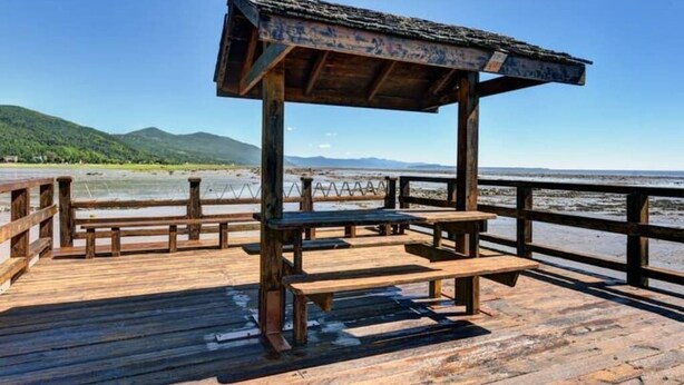 A wooden deck with benches and a covered area overlooking a beach and mountains. - Hôtel à la maison - La Maison Elsa, fleuve et ski (Petite-Rivière-Saint-François)