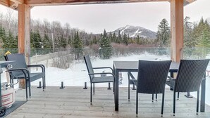 Una terraza de madera con barandilla de vidrio, mobiliario exterior y vistas a una montaña nevada.