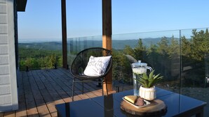 Ein Balkon mit Glastrennwand, einer Holzdielenfläche, einem schwarzen Stuhl, einem Tisch mit Pflanze und einem Getränk sowie einer reizvollen Aussicht auf die Landschaft.