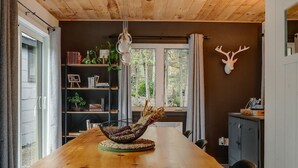 Ein Holzesstisch mit einem Korbbasket als Mittelpunkt, ein Bücherregal mit Büchern und Pflanzen, eine Hirschgeweih-Wanddekoration und ein Fenster mit Blick auf Bäume.