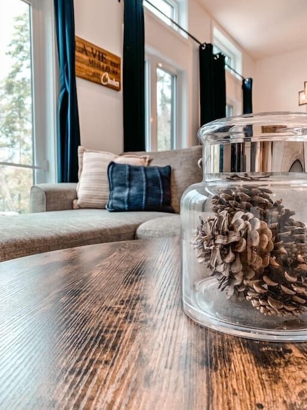 Un frasco de vidrio transparente con piñas sobre una mesa de madera, un sofá con cojines y una ventana con cortinas en una habitación.