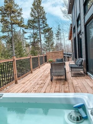 Una terraza de madera con un jacuzzi, dos sillas de mimbre y vistas a árboles.