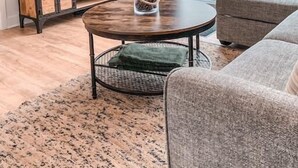 A modern living room with a flat-screen TV, a wooden entertainment unit, a round coffee table, and a large window with a view of trees. - Hôtel à la maison - Chalet la Carpe, Spa et Lac (Chertsey)