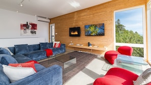 A modern living room with a fireplace, a sofa set, a coffee table, and a large window offering a view of the outdoors. - Hôtel à la maison - Panache, spa et fleuve (Baie-Saint-Paul)