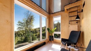 Una habitación moderna con ventanas amplias, una silla negra, una mesita con vasos y una planta en maceta.