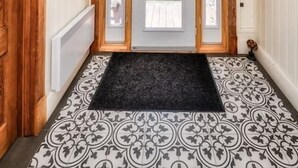 A hallway with a wooden door, a black doormat, patterned floor tiles, and a ceiling light fixture.