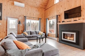 Un salon chaleureux avec une cheminée, des canapés gris et de grandes fenêtres donnant sur la forêt.