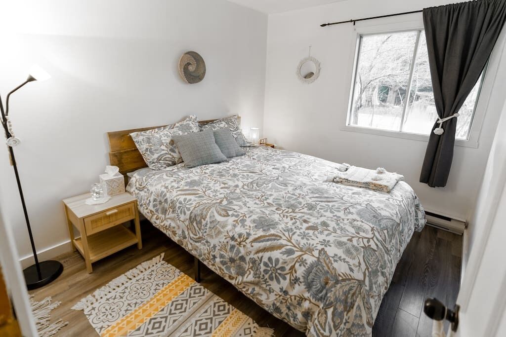 Une chambre à coucher avec un lit, une table de chevet, une lampe, une fenêtre et des rideaux.
