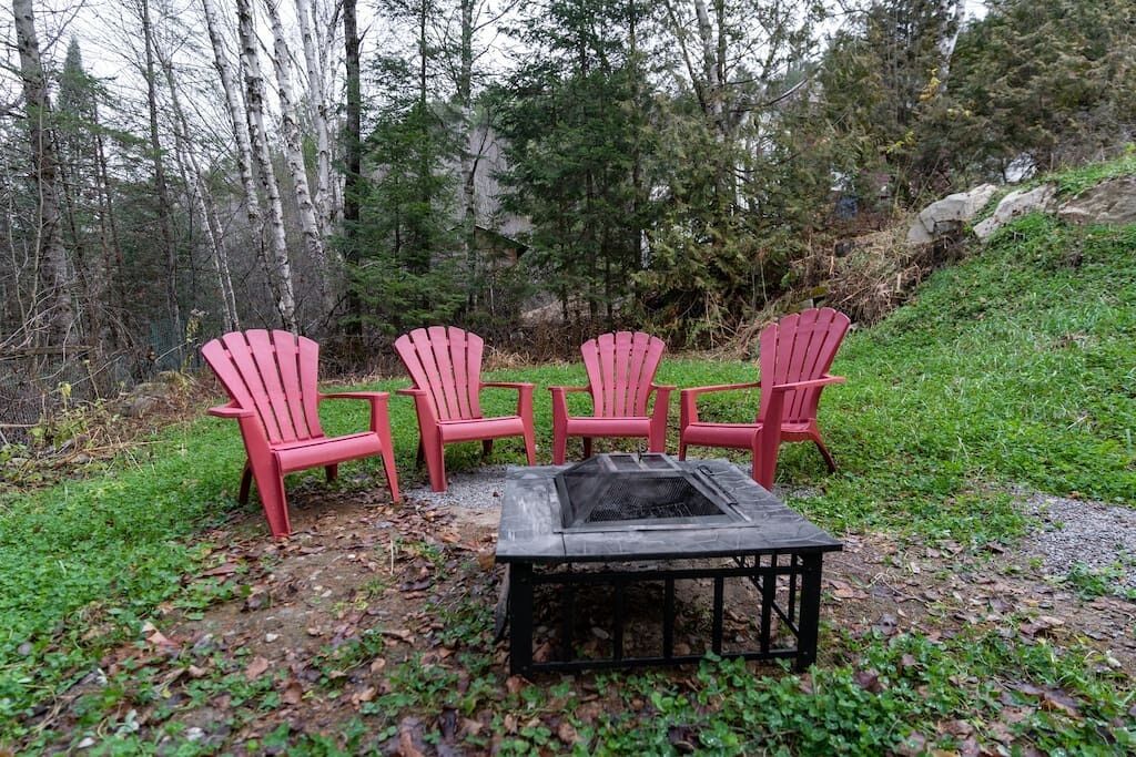 Un hogar con fuego rodeado por cuatro sillas Adirondack rojas en un área boscosa.