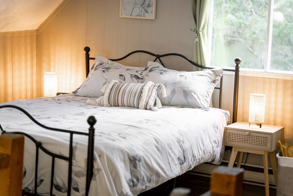 Un dormitorio con cama, dos mesitas de noche, una ventana con cortinas e imagen colgada en la pared.