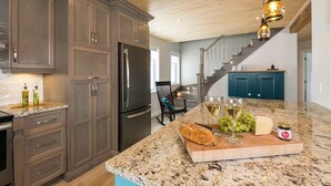 Una cocina con armarios oscuros, un refrigerador de acero inoxidable, una encimera de granito y una isla de madera con una barra de pan, uvas y una copa de vino.