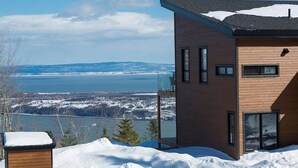 A modern two-story building with large windows and a view of a lake and mountains. - Hôtel à la maison - L'Horizon, spa et fleuve (Les Éboulements)