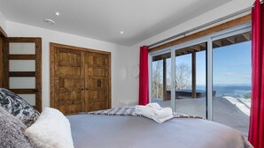 A bedroom with a large bed, a wooden wardrobe, and a sliding glass door leading to a balcony with a view. - Hôtel à la maison - L'Horizon, spa et fleuve (Les Éboulements)