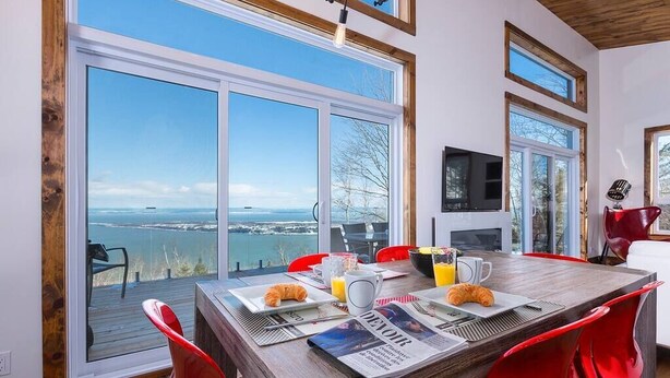 A dining area with a large table, red chairs, breakfast items, and a view of a body of water through large windows. - Hôtel à la maison - L'Horizon, spa et fleuve (Les Éboulements)