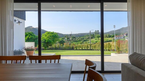 Una sala de estar moderna con un amplio ventanal corredera que da a un balcón, muebles de madera y vistas a un paisaje verde.