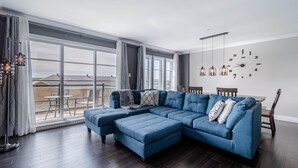 Una sala de estar moderna con un amplio sofá modular, una mesa de comedor con sillas y vistas de la ciudad a través de grandes ventanales.