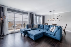 Una sala de estar moderna con un amplio sofá modular, una mesa de comedor con sillas y vistas de la ciudad a través de grandes ventanales.