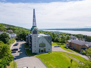 A church with a tall steeple and a stone building with a green roof. There is a parking lot with several cars and a grassy area in front.