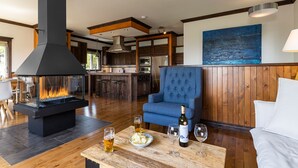 A modern living room with a fireplace, a blue armchair, a wooden coffee table with drinks, and a kitchen in the background. - Hôtel à la maison - L'Escale, Spa et Fleuve (Les Éboulements)