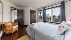A bedroom with a large window, a bed with striped bedding, a wicker chair, and a wooden door. - Hôtel à la maison - L'Escale, Spa et Fleuve (Les Éboulements)