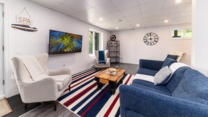 A modern living room with a sofa, armchair, and a coffee table. There is a painting on the wall, a clock, and a sign that reads "SPA". - Hôtel à la maison - L'Escale, Spa et Fleuve (Les Éboulements)