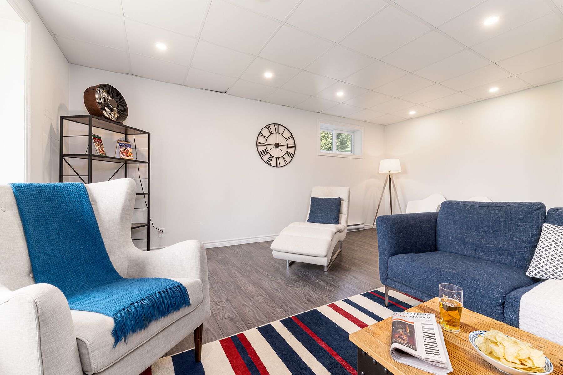 A modern living room with a white sofa, a blue armchair, a small white chair, a striped rug, a wooden coffee table with a newspaper and a glass of beer, and a bookshelf with a clock and a lamp.