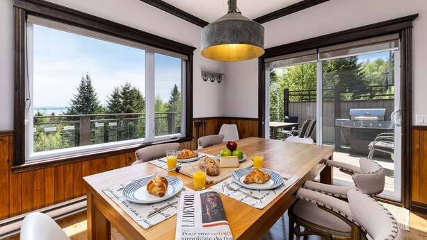 A dining area with a wooden table, chairs, and a view of the outdoors. - Hôtel à la maison - L'Escale, Spa et Fleuve (Les Éboulements)
