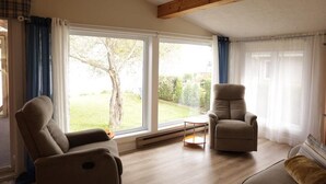 Een kamer met twee beige fauteuils, een kleine ronde tafel en een groot raam met gordijnen.