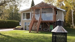 Een huis met twee verdiepingen, met een houten trap naar een balkon, een schoorsteen en een metalen vogelbad in de tuin.