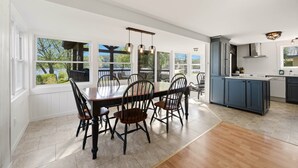 Een ruime eethoek met een grote tafel en stoelen, een keuken met blauwe kasten, en grote ramen met uitzicht op groen.