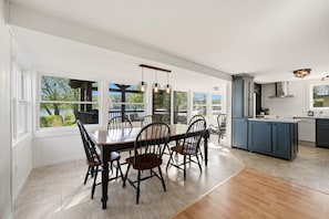 Un vaste espace de salle à manger avec une grande table et des chaises, une cuisine dotée de meubles bleus et de grandes fenêtres offrant une vue sur la verdure.