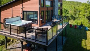 Ett modernt hus med ett bubbelbadkar inbyggt i en glasstruktur, utomhusmöbler och en terrass med utsikt över ett skogsområde.