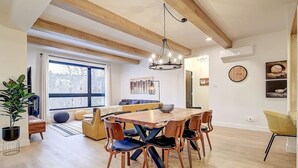 A spacious dining area with a wooden table and chairs, a chandelier, and a large window providing natural light.