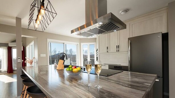 A modern kitchen with a large island, stainless steel appliances, and a view of the outdoors. - Hôtel à la maison - Colombe du Cap, foyer et spa (Baie-Saint-Paul)