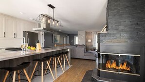 A modern kitchen with a central island, bar stools, a built-in fireplace, and a stone accent wall. - Hôtel à la maison - Colombe du Cap, foyer et spa (Baie-Saint-Paul)