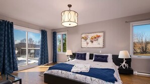 A bedroom with a large bed, two bedside tables with lamps, a window with curtains, and a painting on the wall. - Hôtel à la maison - Colombe du Cap, foyer et spa (Baie-Saint-Paul)