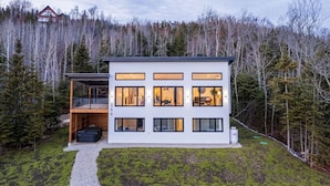 Una casa moderna de dos pisos con ventanas amplias y un porche cubierto, rodeada de un área boscosa.